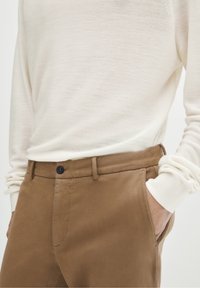 Osoba ubrana w kremową koszulę z długim rękawem włożoną w brązowe spodnie, z jedną ręką swobodnie w kieszeni, na jednolitym tle.