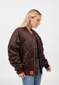Bruine satijnen bomberjack met een diamantquilt-patroon, geribde manchetten en een oranje label aan de zoom. Gemaakt over een zwarte top en blauwe jeans.