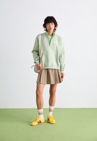 Sweatshirt zippé vert menthe avec une poche avant, jupe beige plissée, baskets jaunes et chaussettes rayées. Matière en mélange de coton, design décontracté.