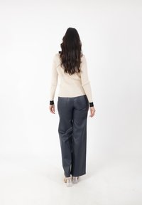 Suéter de punto acanalado beige con puños negros, combinado con pantalones de cuero oscuros de pierna ancha. Modelo de espaldas, con el cabello largo y ondulado visible.