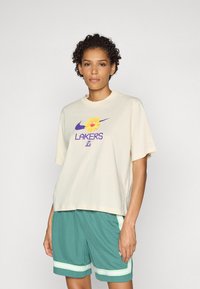 Kremo spalvos marškinėliai su purpurine "Lakers" logotipu ir gėlių grafika, derinami su žaliais šortais su baltais ir žaliais dryžiais.