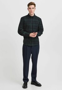 Cămașă în carouri de verde închis și negru, cu închidere prin butoane și guler, asortată cu pantaloni negri croiți și pantofi negri.