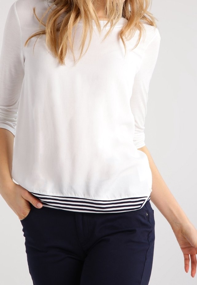 Femme aux longs cheveux blonds portant un haut blanc à manches longues avec un ourlet rayé et un pantalon bleu foncé, debout, une main dans la poche.