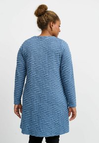 Långärmad, texturerad klänning i mjukt, blått tyg med ett vågigt mönster. Har en rund halsringning och når till mitten av låret.