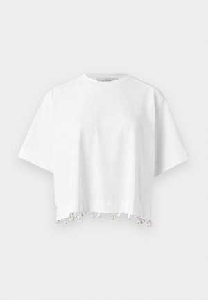 Žena má na sebe čierne obdĺžnikové slnečné okuliare, biely krátky top s lemom s brmbolcami, sivé široké krátke nohavice po kolená a čierne baleríny so zadným remienkom.