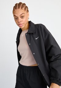 Juoda vėjo neperpučiama striukė su smailiu apykakle ir spaudėmis, dėvima virš šviesiai rudos palaidinės, derinama su juodomis elastingomis kelnėmis. Matomi Nike logotipai.