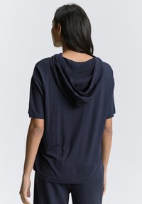 Mørkeblå hoodie i blødt stof, med afslappet pasform, korte ærmer og hætte med snoretræk. Sømligheder synlige bagpå.