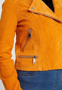 Oranje suède motorjack met een asymmetrische rits, zilveren hardware en twee ritszakken. Zachte textuur en een aansluitend ontwerp.