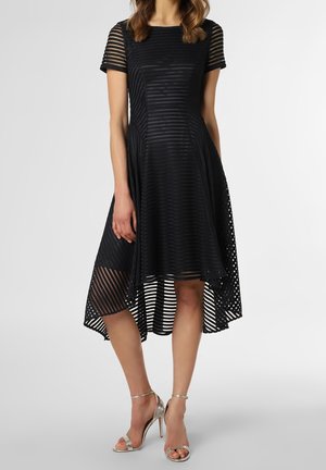 Frau trägt ein schwarzes High-Low-Kleid mit durchscheinenden horizontalen Streifen und kurzen Ärmeln, kombiniert mit silbernen Sandalen mit hohem Absatz.