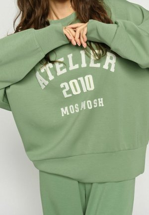 Persoon draagt losse groene trui en broek met witte letters "ATELIER 2010 MOS MOSH" aan de voorkant, handen geklemd bij de borst.