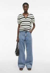 Camisa de polo de punto a rayas en blanco y negro con escote en V, combinada con unos vaqueros de pierna ancha en azul. La modelo sostiene un bolso negro y lleva zapatos de tacón abiertos.