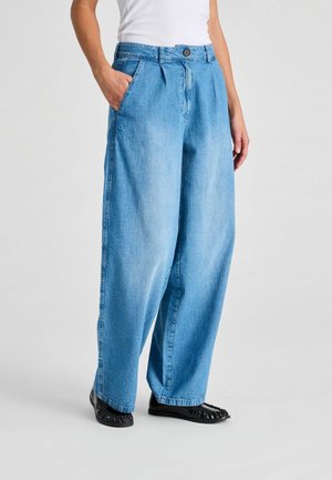 Kobieta ubrana w jasnoniebieskie jeansy o szerokich nogawkach, biały top i czarne plecione płaskie buty, stojąca z jedną ręką w kieszeni.