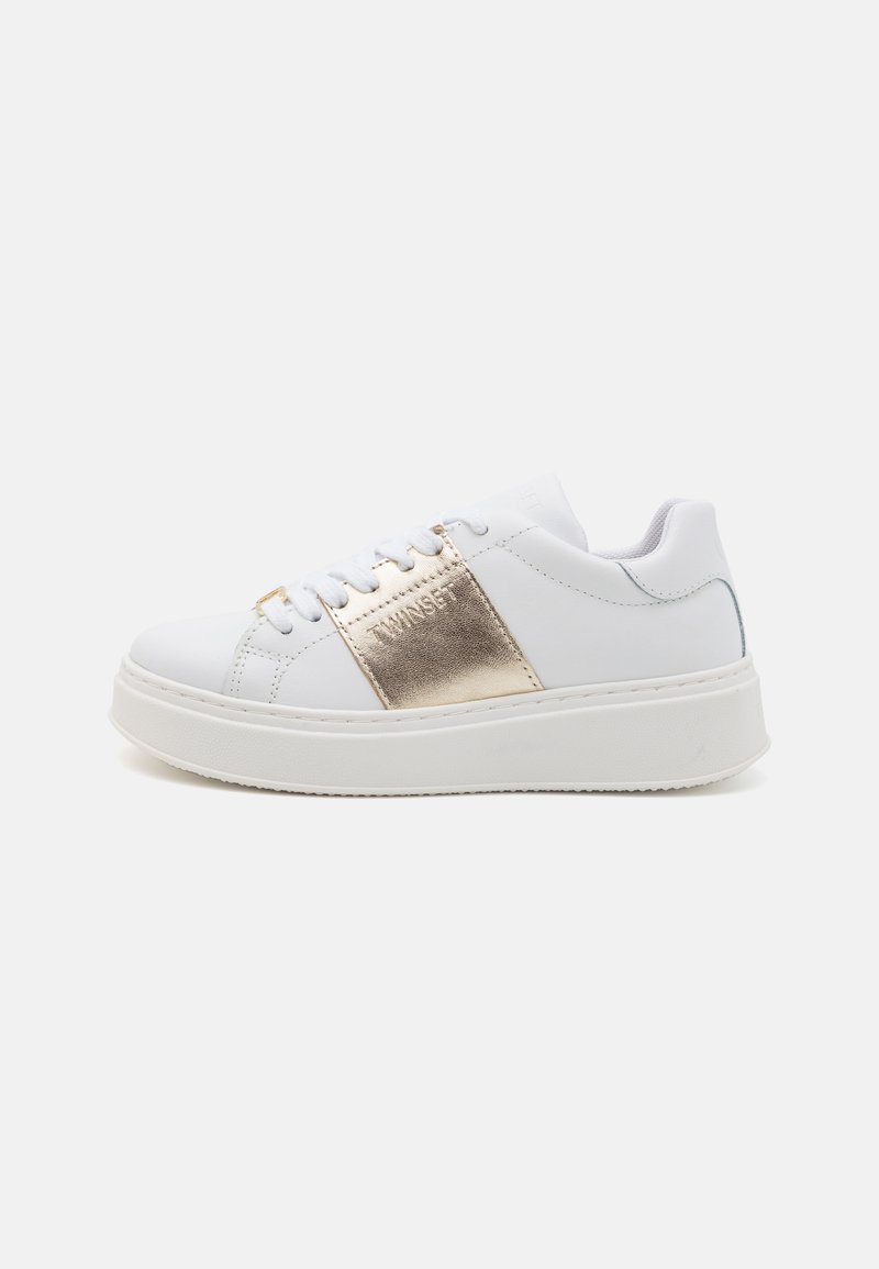 Baskets blanches avec une semelle surélevée, présentant une bande métallique dorée et un logo sur le côté. Texture lisse et fermeture à lacets.