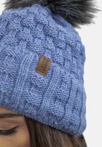 Căciulă tricotată albastră cu un model texturat și un pom-pom din blană sintetică neagră. O etichetă mică din piele maro este coasă pe față.
