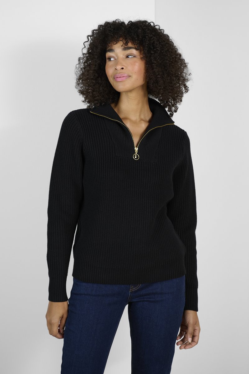 Pull en maille côtelée noire avec un col zippé et des détails en métal doré, doté de manches longues et d'une coupe décontractée. Porté avec un jean bleu.