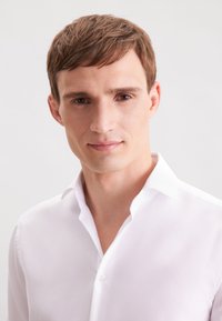 Camicia bianca con colletto strutturato, caratterizzata da una trama sottile e un tessuto liscio. La camicia è indossata da un modello maschile con i capelli corti.