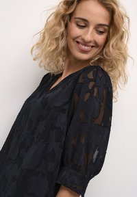 Cappotto nero con sovrapposizione in pizzo floreale trasparente, maniche a tre quarti e vestibilità morbida. I dettagli texture arricchiscono la profondità del tessuto.