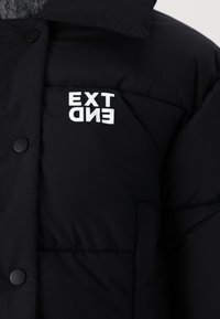 Sort pufferjakke med høj krave, prydet med et fremtrædende hvidt "EXT" og "END" logo, trykknapper og et struktureret stof.