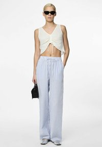 Witte crop top met een geplooid ontwerp, gecombineerd met lichtblauwe en witte gestreepte wijde broek. Model houdt een kleine zwarte tas vast.