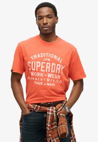 Orange bomulds T-shirt med hvidt print, der siger "SUPERDRY WORK-WEAR." Kombineret med en plaid flannel-skjorte bundet om taljen, blå jeans og et ur.