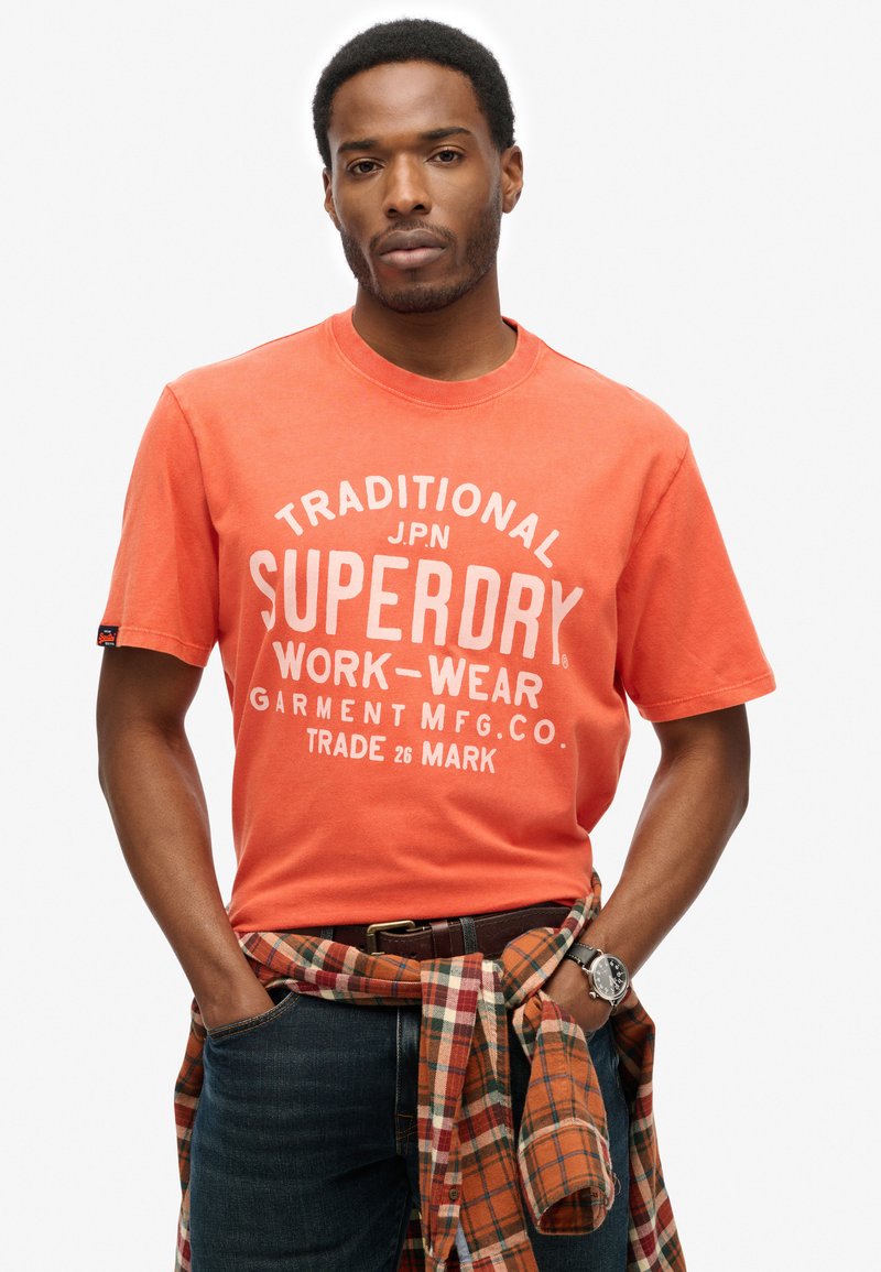 Orange bomulds T-shirt med hvidt print, der siger "SUPERDRY WORK-WEAR." Kombineret med en plaid flannel-skjorte bundet om taljen, blå jeans og et ur.