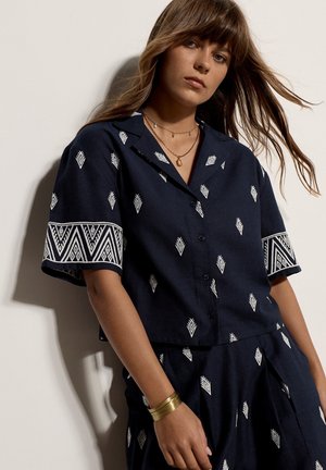 Jeune femme aux longs cheveux porte un ensemble chemise à manches courtes et jupe bleu marine avec des motifs géométriques blancs ainsi que des bijoux en or, debout devant un mur clair.