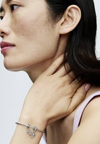 Il bracciale argento presenta un design intrecciato con chiusure a gancio e un accento a forma di cuore. Il soggetto ha un tono di pelle neutro con capelli scuri di lunghezza media.