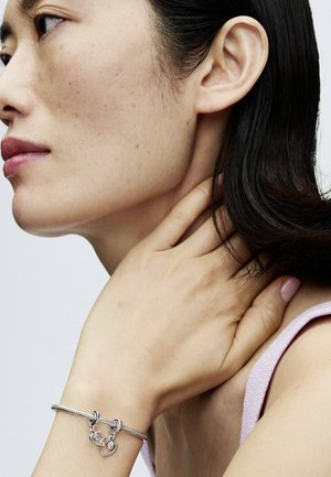 Le bracelet en argent présente un design tressé avec des fermoirs à crochet et un accent en forme de cœur. Le sujet a un teint neutre et des cheveux foncés de longueur moyenne.