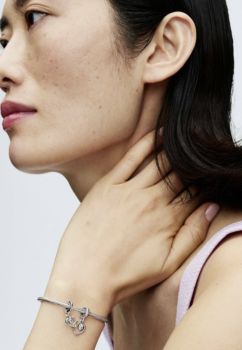 Il bracciale argento presenta un design intrecciato con chiusure a gancio e un accento a forma di cuore. Il soggetto ha un tono di pelle neutro con capelli scuri di lunghezza media.