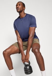 Navy athletic shirt with short sleeves and light olive shorts, holding a black kettlebell, on a plain white background.