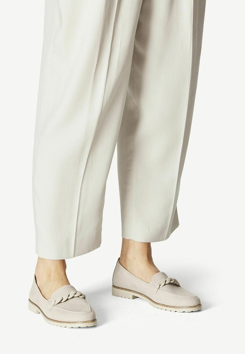 Chaussures slip-on beiges avec une accentuation tressée, présentant un dessus texturé et une semelle en caoutchouc de couleur claire. Associées à un pantalon large couleur crème.