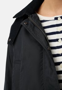 Zwarte jas met een corduroy kraag, ritsluiting, zijzak en een gladde textuur. Daaronder is een gestreept shirt met knopen zichtbaar.