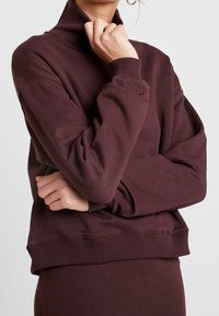 Mujer vestida con un sudadera de cuello alto en un tono mauve oscuro y una falda a juego, con los brazos cruzados, frente a un fondo liso y claro.