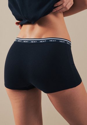 Osoba zdvíhajúca námornícky modrý top, oblečená v čiernych priliehajúcich boxerkoch s pásom s nápisom „NEXT“ bielymi písmenami.