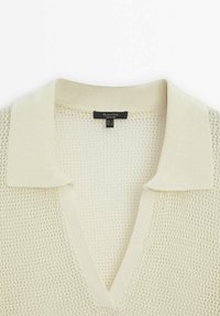 Polo en maille ajourée de couleur crème avec col en V et col côtelé, doté d'une étiquette noire indiquant la taille et la marque au niveau du col.