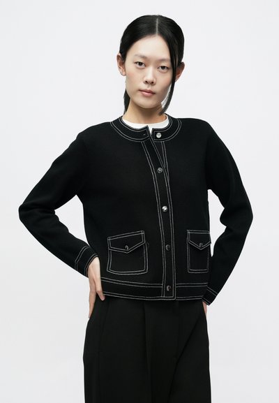 Fekete kardigán kontrasztos fehér varrással, kerek nyakkal, elöl gombos záródással és két felvarrt zsebbel az elején. Puha tapintású anyag.