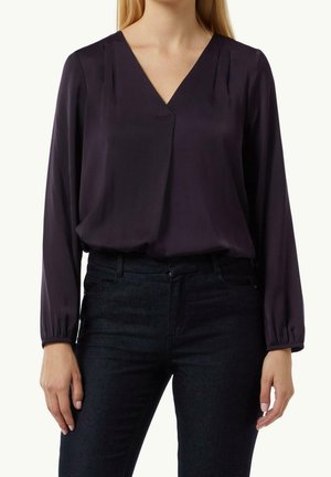Kvinde iført en mørkelilla V-hals bluse med lange ærmer, der er stoppet ned i højtaljede sorte jeans, står med armene afslappet ned langs siderne.
