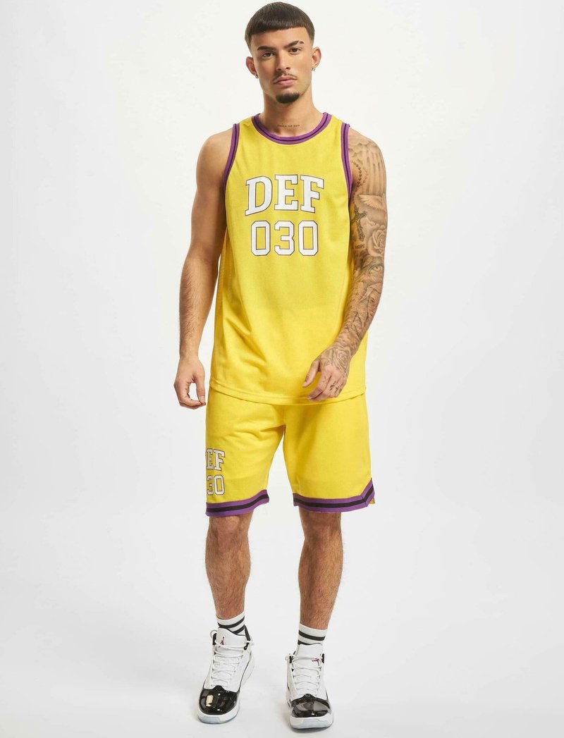 Gelbes Tanktop und Shorts-Set mit lila Akzenten und weißer Schrift "DEF 030." Der Stoff scheint atmungsaktiv und strukturiert zu sein. Die Sneakers sind schwarz und weiß.