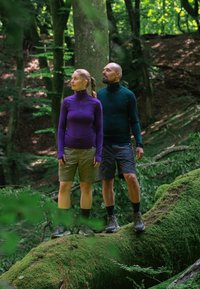 Zwei Personen stehen auf einem moosbedeckten Baumstamm im Wald. Eine trägt ein lila langärmliges Oberteil und khakifarbene Shorts; die andere trägt ein dunkelgrünes Oberteil und Shorts.
