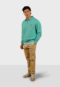 Pullover turchese con colletto a zip, caratterizzato da un patch ricamato con logo. Abbinato a pantaloni beige e sneakers grigie con lacci bianchi.