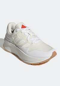 Chaussure de sport légère en mesh blanc et matériaux synthétiques. Comprend trois bandes blanches, un détail rouge, et une semelle extérieure en caoutchouc gum.