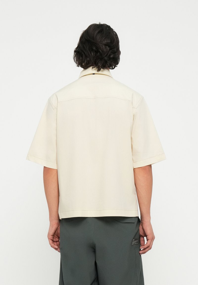 Camicia beige a maniche corte con orlo dritto, colletto a punta e dettagli di cucitura sottili; indossata con pantaloni verde scuro, che mostrano un design minimalista.