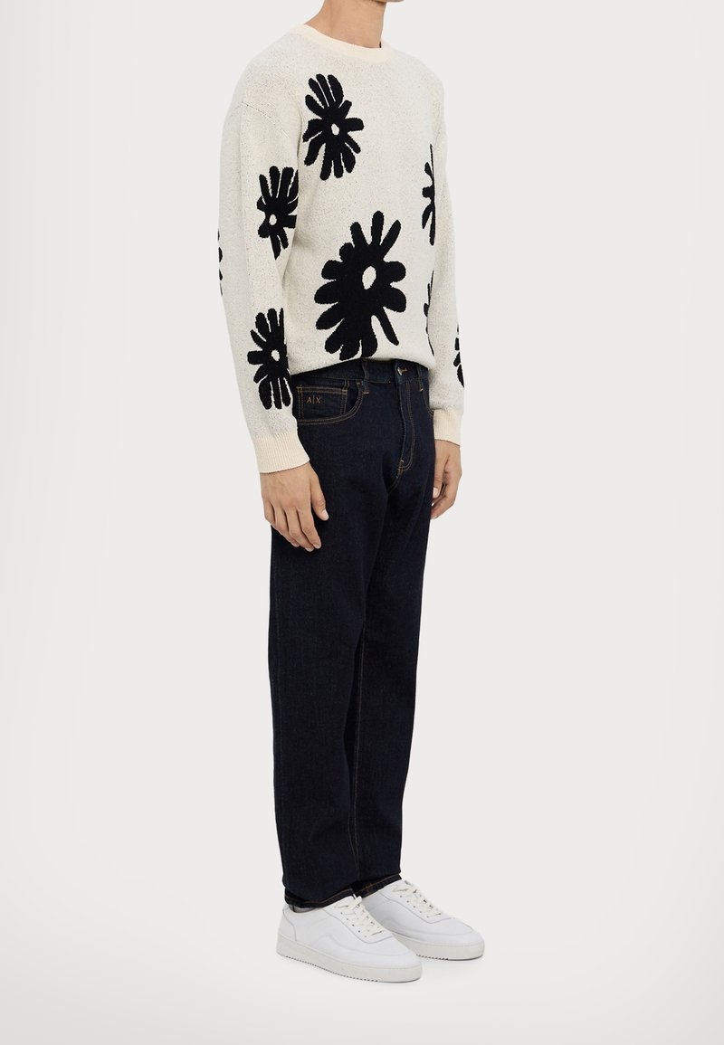 Personne portant un pull clair avec de grands motifs floraux noirs, un jean foncé et des baskets blanches, debout devant un fond uni.