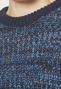 Námořnický modrý pletený svetr s vroubkovaným límcem. Obsahuje texturovaný vzor propletených tmavě a světle modrých nití.