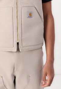 Beige vest med gullglidelås og to frontlommer, med et Carhartt-logo-påsydd merke. Beige bukser med gullfargede lommer.