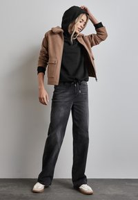 Chaqueta de forro polar marrón con un cuello de borreguito, sudadera negra debajo, jeans negros y zapatillas blancas, en una postura casual.