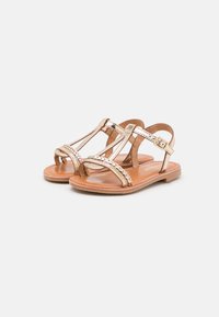 Sandali metallici dorati con design a T-strap. Presentano dettagli intrecciati e una fibbia regolabile. Suola marrone, profilo piatto e texture liscia.