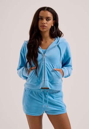 Kvinde med langt mørkt hår iført lyseblå velour-hoodie med lynlås og matchende shorts med snor, hænderne i hoodie-lommerne, neutral baggrund.