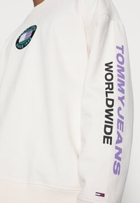 Felpa color crema con logo circolare ricamato e testo "TOMMY JEANS WORLDWIDE" in nero e viola sulle maniche.