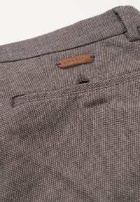 Braun strukturiertes Gewebe mit einem kleinen Patch-Label, auf dem "bugatti" steht. Verfügt über eine Gesäßtasche mit einem Knopf und Steppnähten.